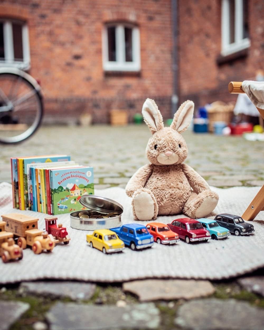 Auf einer hellen Decke im Hinterhof ausgelegtes Kinderspielzeug: eine Reihe bunter Modellautos, eine Holzeisenbahn, ein Plüschhase, aufgereihte Bilderbücher und eine offene Blechdose mit Münzen, dahinter eine Backsteinfassade.