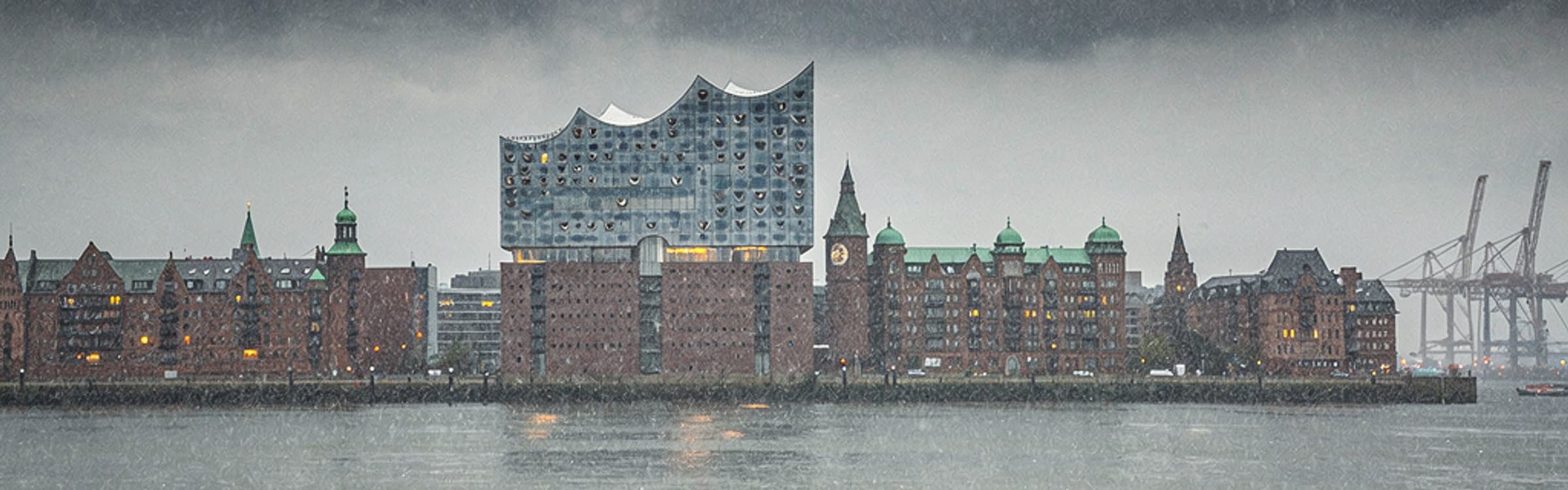 Die Hamburger Hafensilhouette bei starkem Regen, von der Elbe aus gesehen: Elbphilharmonie, Backsteinspeicher mit gruenen Turmspitzen und dahinter Hafenkraene im Dunst.