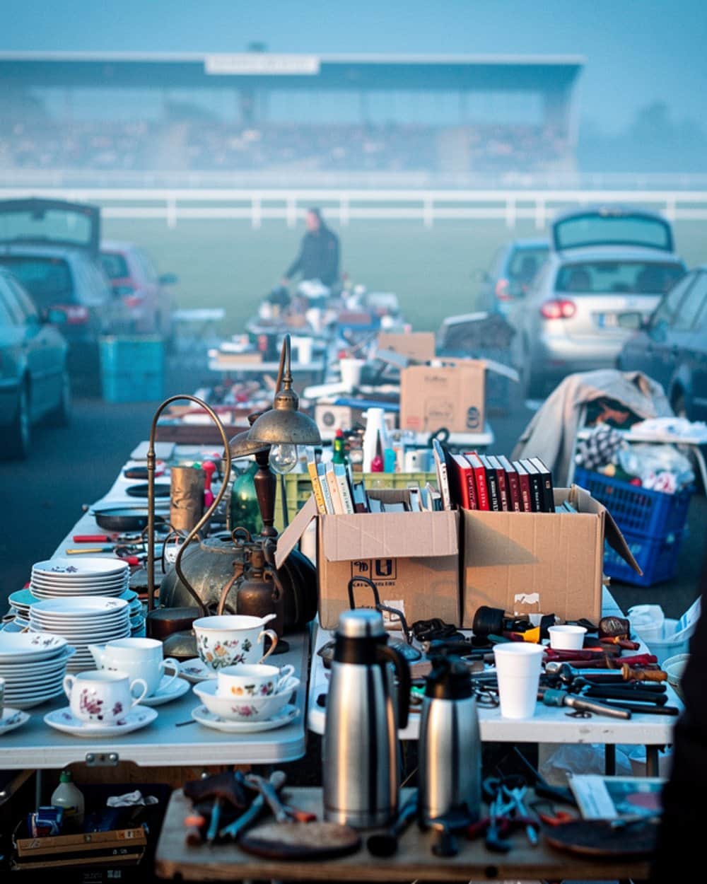 Lange Flohmarkttische mit Geschirr, Büchern und Werkzeug im Morgennebel, dahinter parkende Autos mit offenen Kofferräumen.