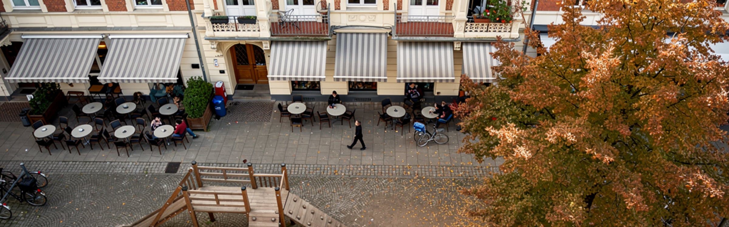 Blick von oben auf einen Hamburger Quartiersplatz am Nachmittag: Gründerzeitfassaden mit gestreiften Markisen und Balkonen, davor runde Cafétische auf dem Pflaster, ein hölzernes Klettergerüst und eine herbstlich gefärbte Kastanie.