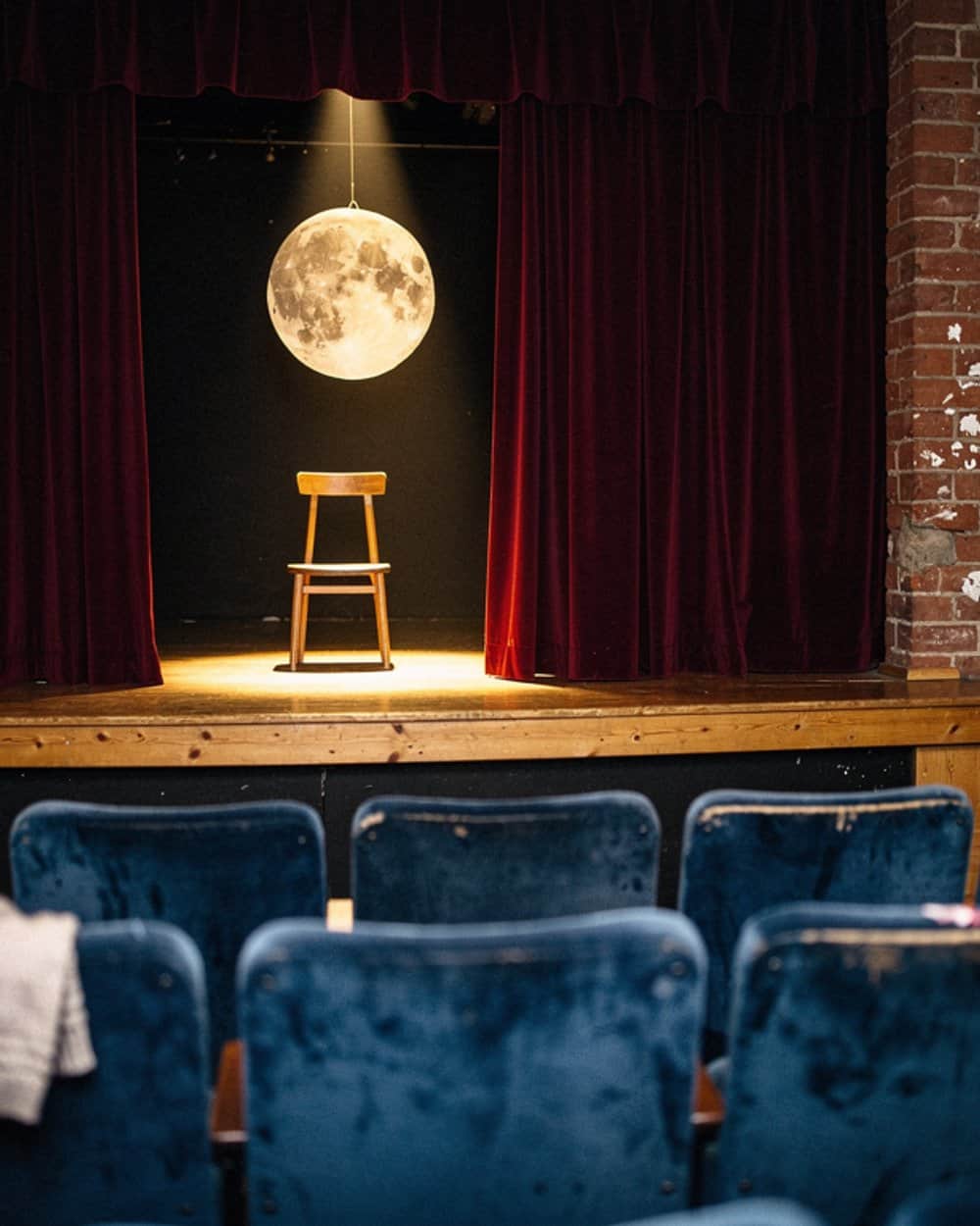 Kleine Theaterbühne mit rotem Samtvorhang, einer leuchtenden Mondkugel und einem Holzstuhl im Scheinwerferkegel, davor blaue Klappsessel.