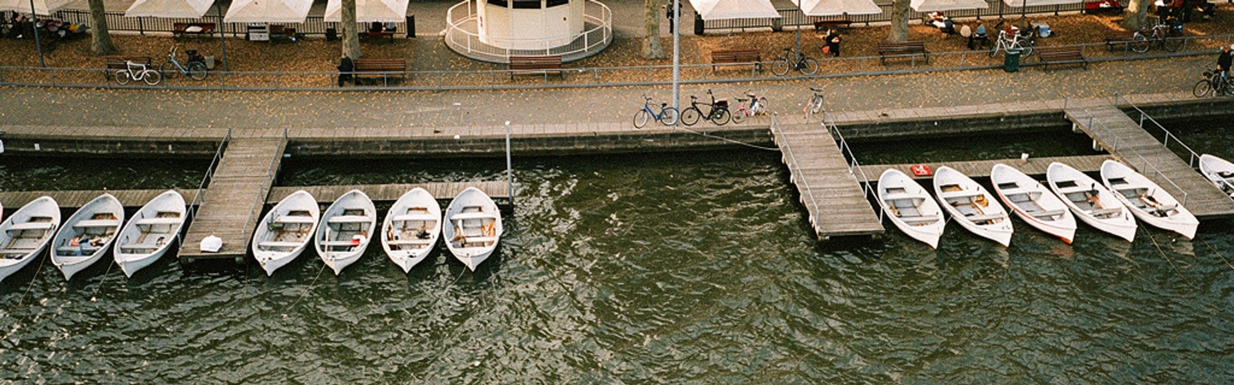 Blick von oben auf die Alsterpromenade in Hamburg an einem Herbstsamstag: weiße Sonnenschirme und ein runder Pavillon unter Platanen, Bänke und abgestellte Fahrräder am Geländer, darunter Holzstege mit aufgereihten weißen Ruderbooten auf dem Wasser.