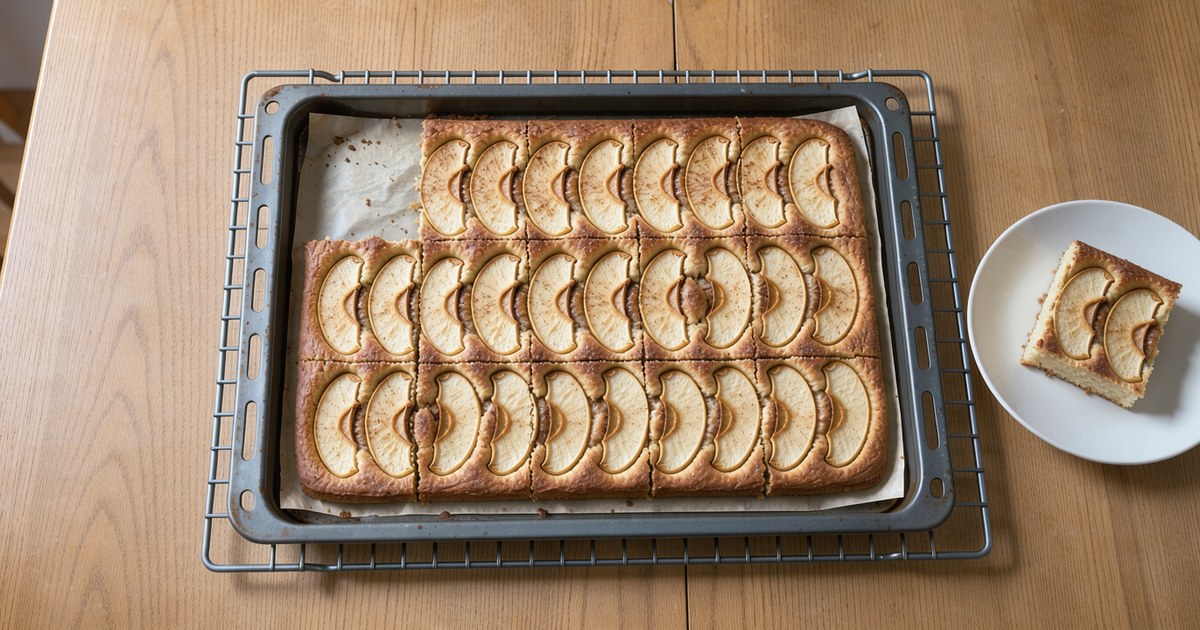 Von schräg oben: ein Backblech mit goldbraunem Apfelkuchen, in Quadrate geschnitten, auf jedem Stück helle, weich gebackene Apfelspalten mit Zimtzucker; davor auf einem kleinen weißen Teller ein einzelnes Stück