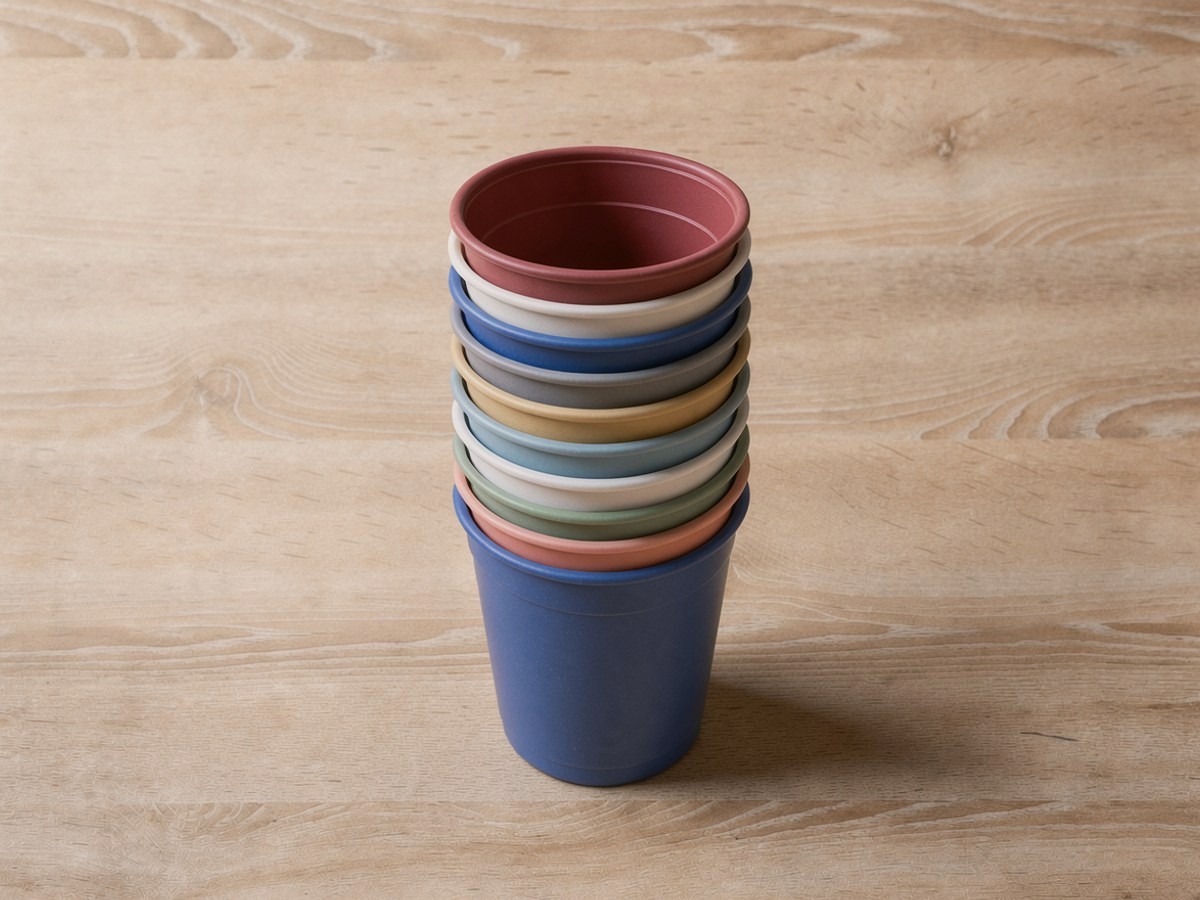 Mehrere einfarbige Becher ohne Aufdruck stecken ineinander und bilden einen hohen schmalen Turm auf hellem Holz, nur die bunten Ränder der einzelnen Becher sind als Streifen zu sehen