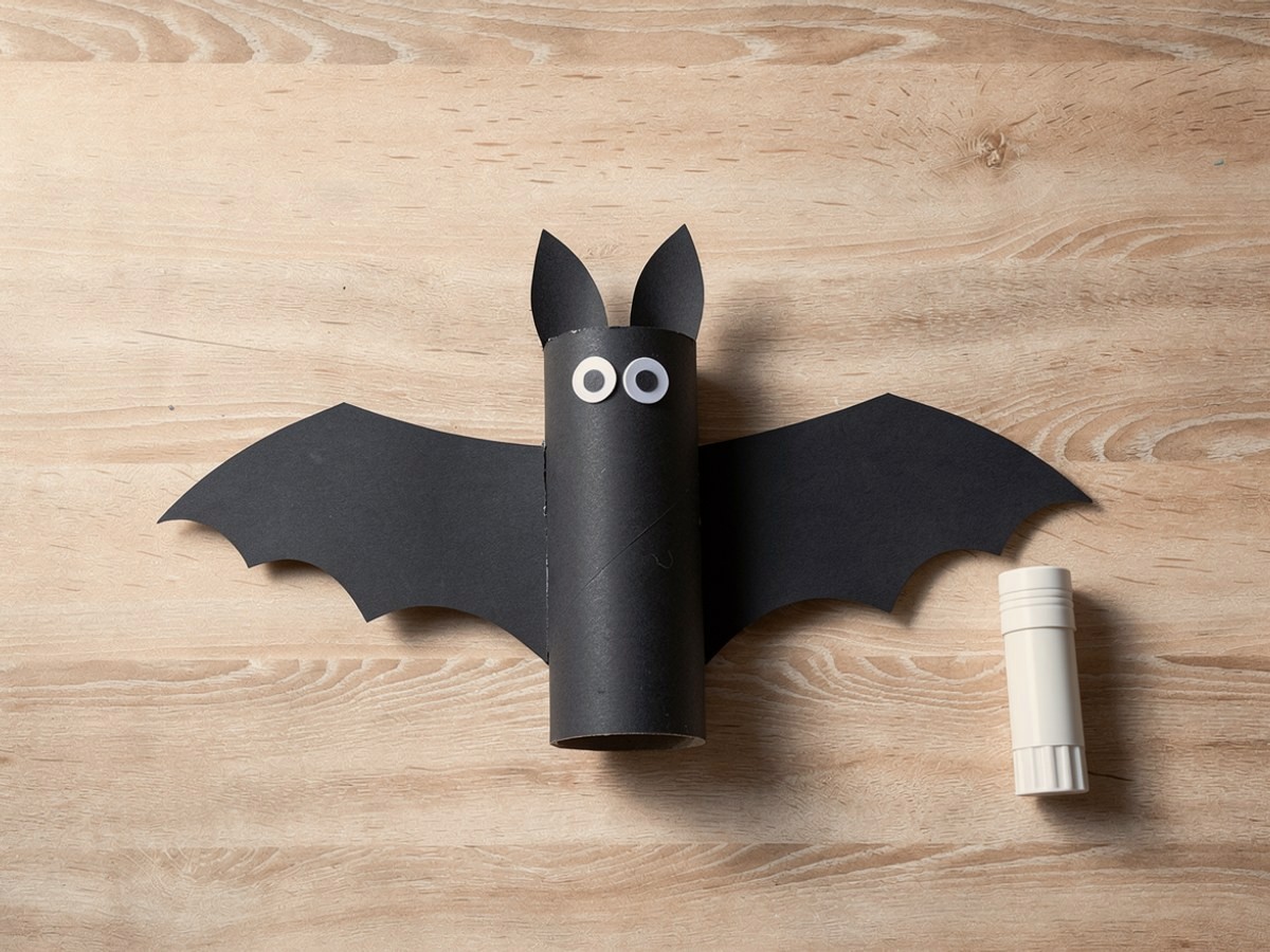 Eine fertige schwarze Fledermaus liegt flach auf hellem Holz, die gezackten Flügel ragen links und rechts heraus, zwei Wackelaugen sitzen unter den eingedrückten Ohren