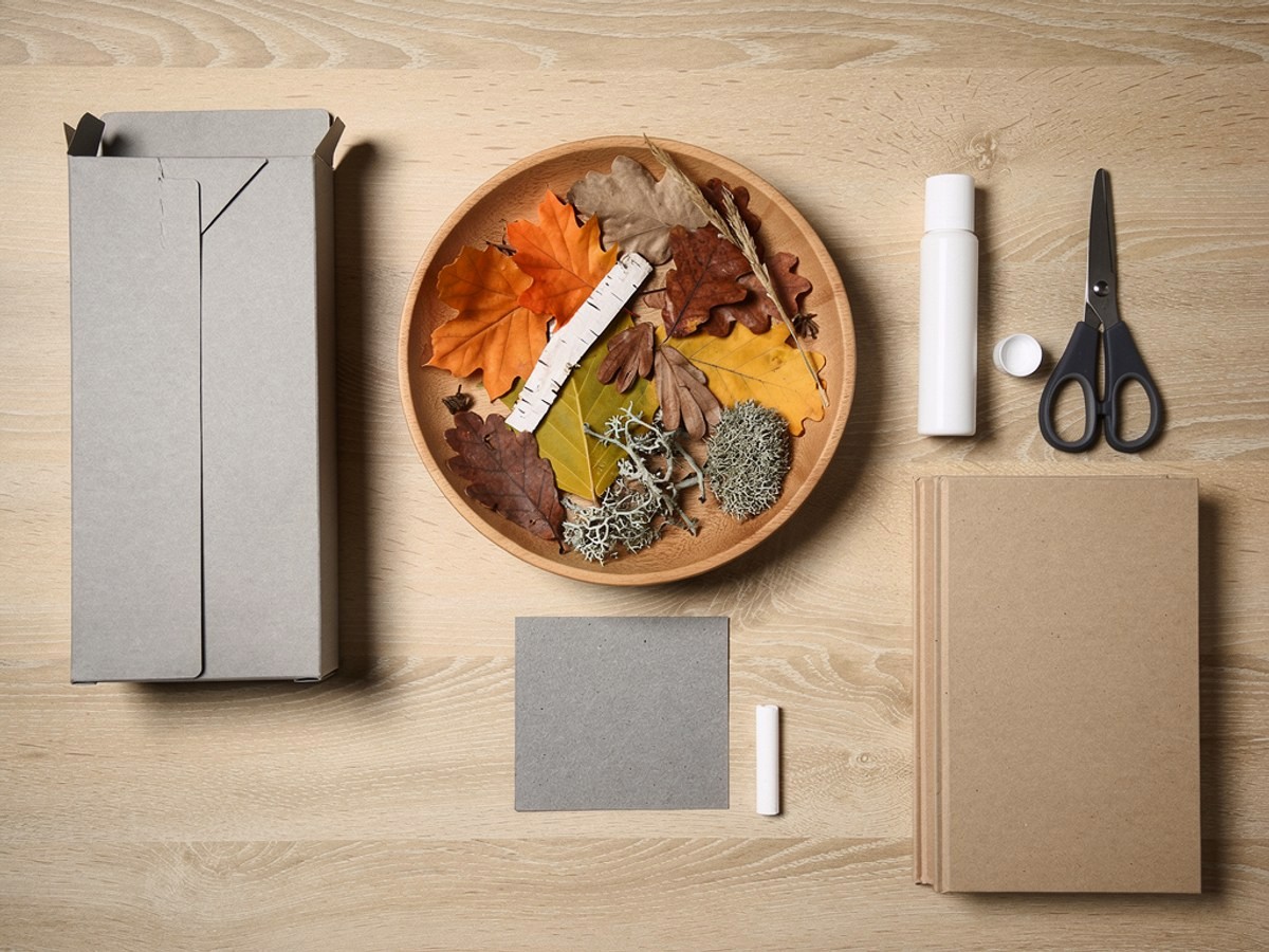 Flatlay auf hellem Holz: eine flache Schale mit bunten Herbstblättern, Grashalmen und Rindenstücken, eine aufgefaltete Pappschachtel, eine Kinderschere, ein Klebestift, ein Stück Kreide und zwei schwere Bücher