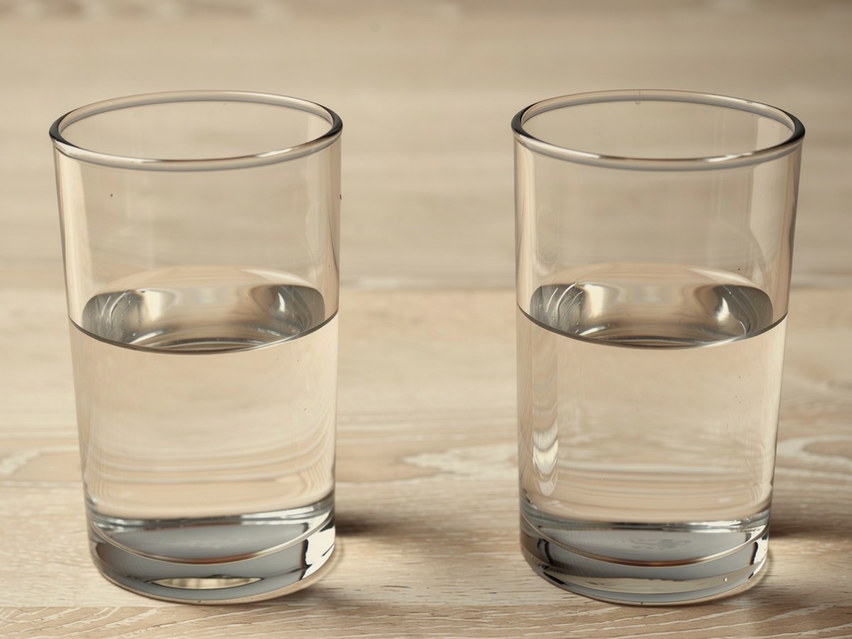 Zwei klare Gläser stehen eine Handbreit auseinander auf hellem Holz, beide gleich hoch mit Wasser gefüllt, daneben der Krug und ein unbemaltes Blatt Küchenpapier
