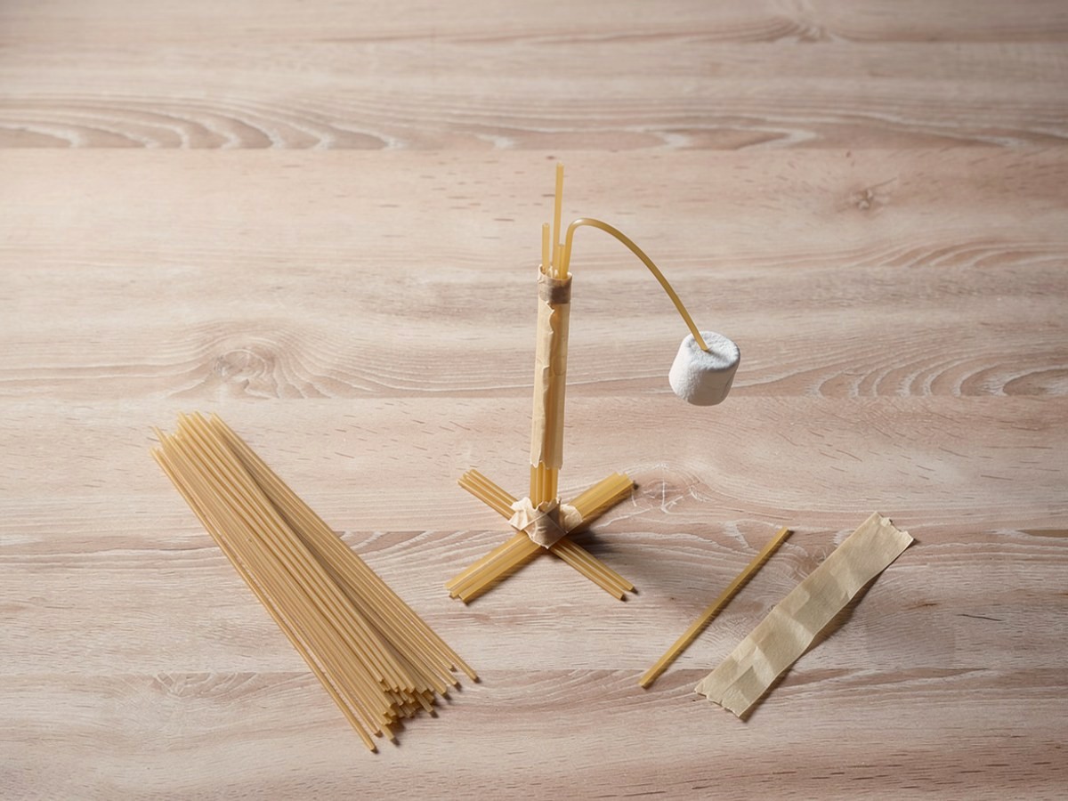 Schräg von oben: eine Säule aus zusammengeklebten Spaghetti ohne Dreiecke auf einem Kreuz aus Spaghetti, oben geht eine einzelne lange Spaghetti weiter und biegt sich im Bogen zur Seite, an ihrer Spitze hängt der Marshmallow auf halber Höhe, daneben liegen lose Spaghetti, ein Rest Klebeband und Schnur