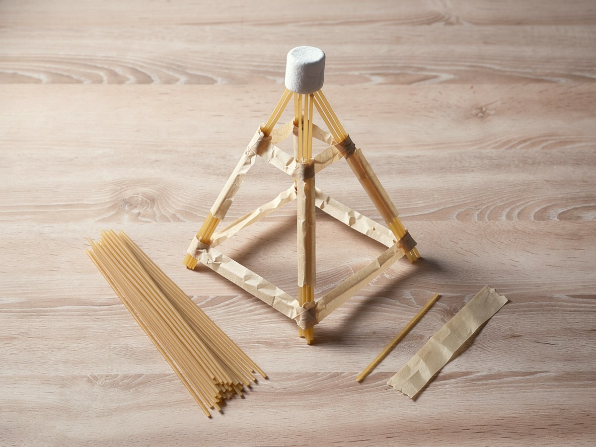 Schräg von oben: ein fertiger Spaghetti-Turm als schlanke Pyramide, die Beine stehen breit auf dem Tisch und laufen oben zusammen, Querstäbe mit Klebeband bilden Dreiecke, ganz oben sitzt der Marshmallow, davor liegen zwei lange Spaghetti, Bruchstücke, ein Rest Klebeband und ein Knäuel Schnur