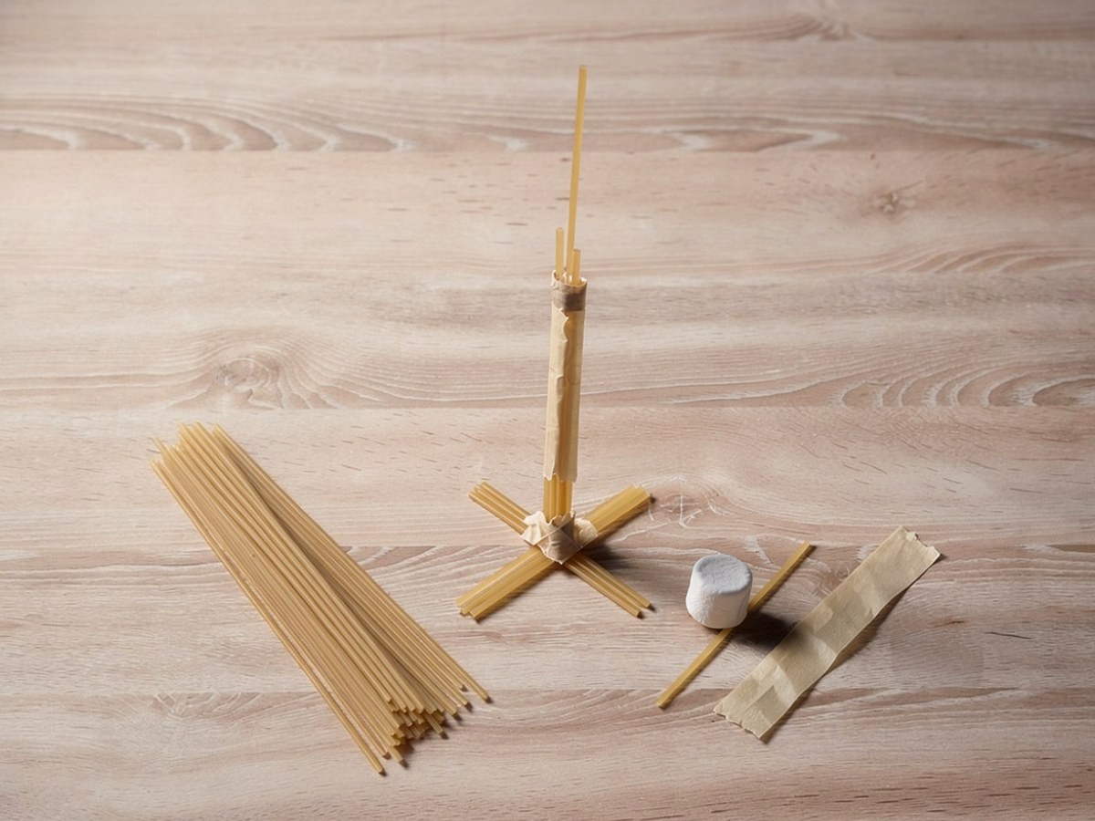 Ein erster, schnell gebauter Turm steht als dünne Säule aus zusammengeklebten Spaghetti auf einem Spaghettikreuz, der Marshmallow liegt noch daneben auf dem Tisch.