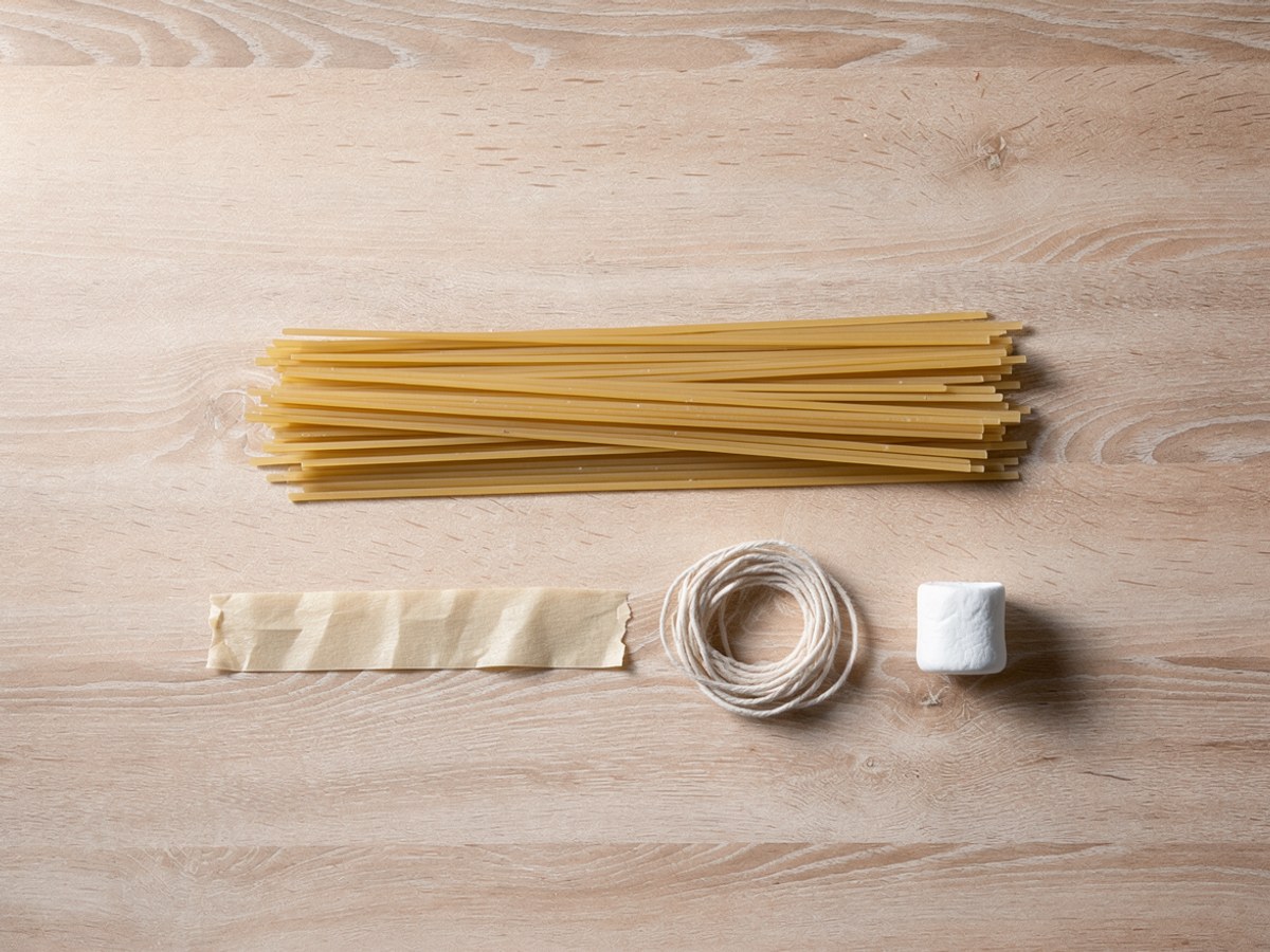 Von oben auf hellem Holz: ein Bündel roher Spaghetti, darunter ein abgerissener Streifen beiges Klebeband, ein kleines Knäuel Naturschnur und ein einzelner weißer Marshmallow