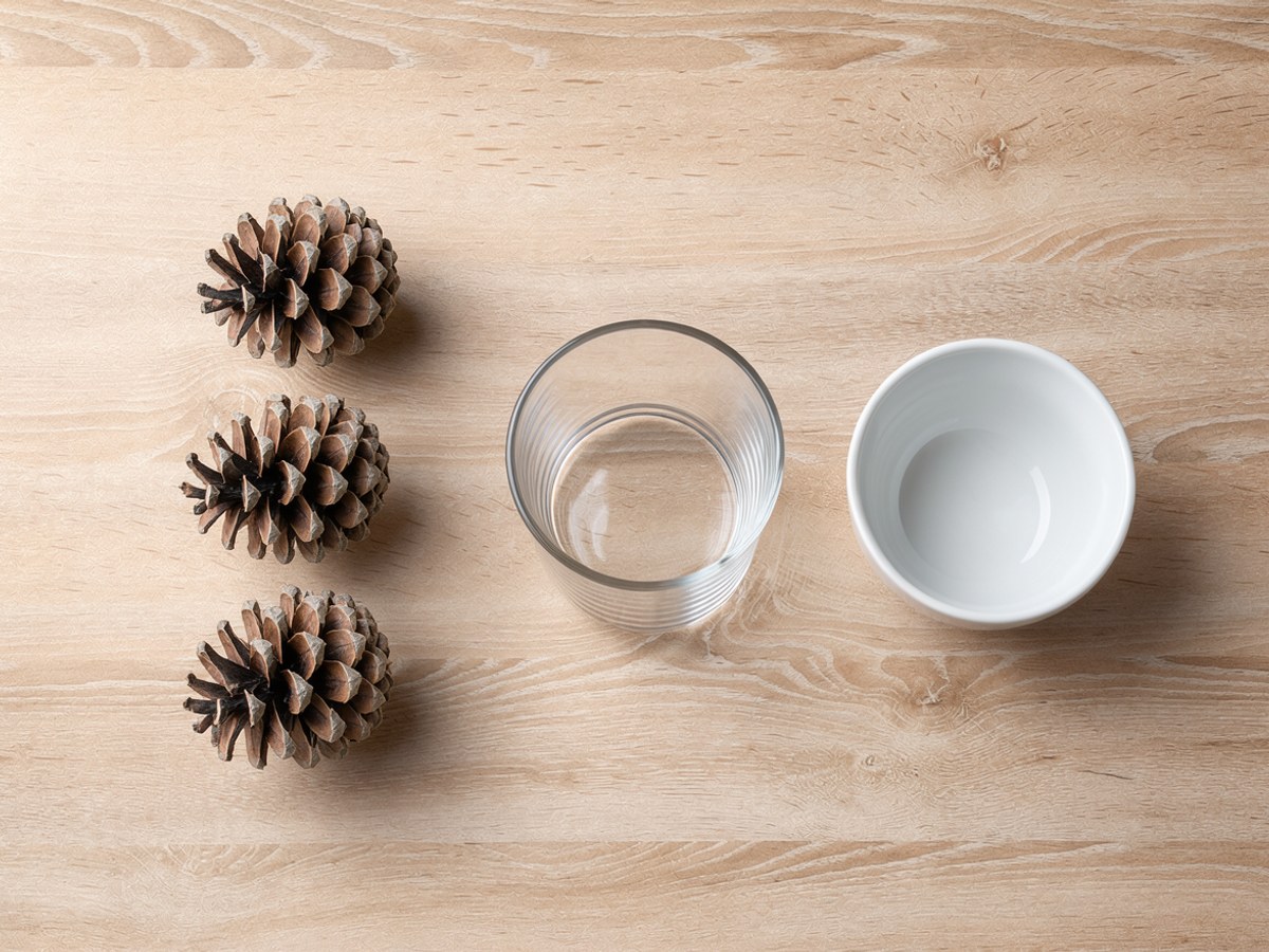 Flatlay auf hellem Holz: drei trockene, geöffnete Kiefernzapfen in einer Reihe, ein hohes leeres Trinkglas und eine kleine weiße Schale